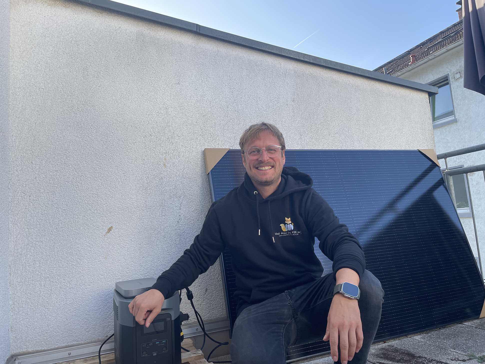 Der Smart-Home-Experte Joe, sitzt neben dem Ecoflow Delta 2 Max Balkonkraftwerk Speicher und dieser ist nachgerüstet und steht angeschlossen an den Powerstream Wechselrichter neben einem Solarpanel.